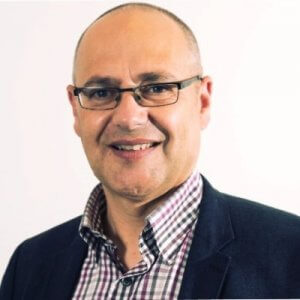 A middle-aged man with glasses and a bald head smiles at the camera, exuding confidence. He is wearing a checkered shirt and a dark blazer, making him look both approachable and professional—a great asset for employers. The background is plain white, highlighting his polished appearance.