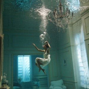 A woman in a flowing white dress is shown underwater in a room with a chandelier, furniture, and large windows. She appears to be gracefully floating or dancing, as if in an ethereal interview with the sea, bubbles rising above her and creating a surreal, dream-like scene.