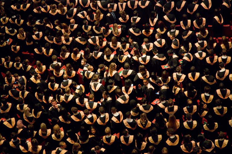 A large group of graduates in caps and gowns sit together in a crowded auditorium, viewed from above. Their gowns are black with white and gold accents. Some hold red folders, signaling their commitment to professional development, and their shiny caps are noticeable in the overhead lighting.