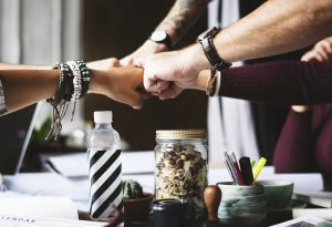 A group of employers joins fists in a gesture of solidarity over a cluttered table with various items such as a jar of mixed nuts, water bottle, and office supplies. All hands display different accessories like bracelets and watches, indicating diverse individuals.