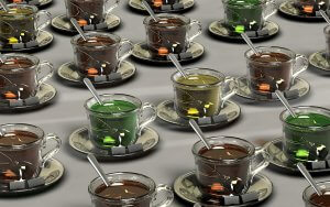 Rows of glass teacups on saucers, each filled with a different-colored liquid ranging from brown to green. A metallic spoon rests in every cup, and a tea bag with a yellow tag is infusing in the liquid. This vibrant display adds a touch of worksite wellness to the simple light gray background.