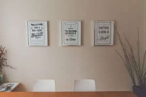 A light-colored wall with three framed motivational posters, each with unique typography. Below the frames, two white chairs and a wooden table create a safe, inviting space. On the left side of the image, there are some magazines and a decorative plant with bare branches.