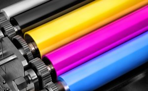 Close-up view of four cylindrical printing rollers lined up in sequence, displaying colors black, yellow, magenta, and cyan from left to right. The rollers are part of a printing press machine with visible gears and metallic parts, essential for maintaining information governance standards in print media.