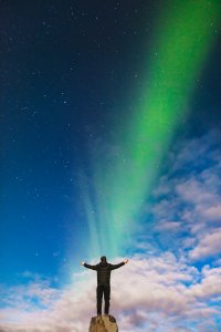 A person stands on a rock with arms outstretched, facing the sky illuminated by vibrant green and blue northern lights. Looking forward, the night is clear with stars visible, and there are some clouds near the horizon.