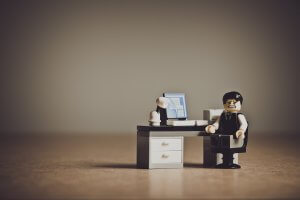 A Lego figure in a black suit and white shirt sits at a small Lego office desk with a computer, desk lamp, and drawers. The figure appears to have a distressed expression, perhaps contemplating the pressures of succession, against a neutral background.