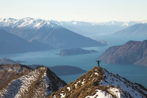 A leader stands triumphantly on top of a snow-capped mountain peak, arms outstretched, overlooking a serene landscape of mountains and a lake with several islands. The sunlight illuminates the vast, rugged terrain under a clear sky.