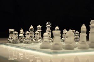 A close-up view of a chessboard with a clear glass and frosted glass chess set. The pieces, resembling poised leaders, are elegantly arranged, reflecting light and creating a pristine and almost ethereal scene. The background is dark, which contrasts sharply with the illuminated chessboard.