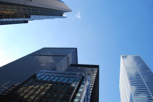 A photo of several modern skyscrapers taken from a low angle, highlighting their varying architectural designs against a clear blue sky. The tall buildings, in perfect succession, feature reflective glass surfaces and sleek lines, emphasizing the urban environment.