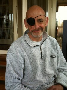 A man with a bald head and a gray beard is sitting on steps, looking forward. He is wearing a light gray pullover and an eye patch over his left eye. The background shows part of a building with glass doors and wooden elements. The man is smiling at the camera.