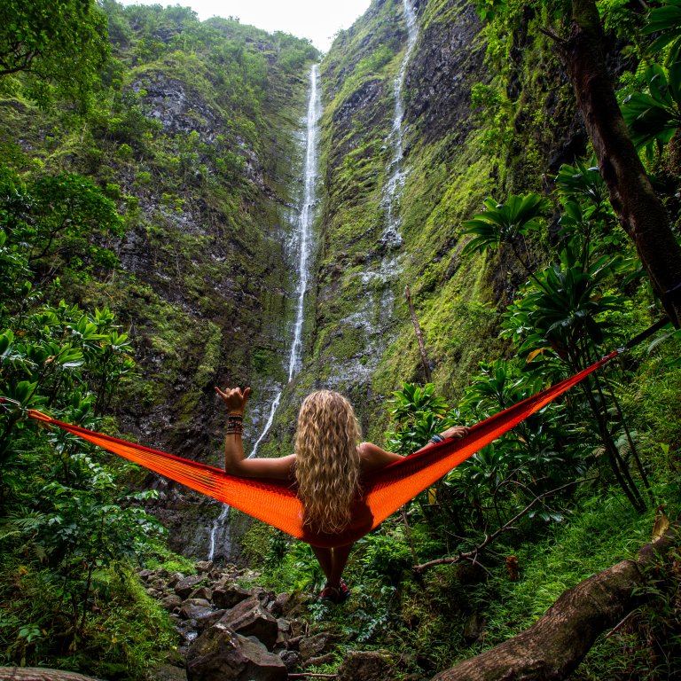 A person with long, wavy blonde hair sits in an orange hammock positioned between two trees in a lush, green forest. Facing tall, cascading waterfalls in the background, surrounded by dense foliage, you find yourself raising one arm in a relaxed pose.