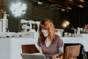 A woman with wavy, red hair is sitting in a cozy cafe, smiling at her laptop. She wears a purple t-shirt, and a bag is placed on a nearby chair. The cafe has a modern interior with a neon sign and coffee machines in the background, creating an inviting atmosphere for the diverse workforce within.