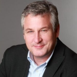 A person with short gray hair and wearing a dark blazer over a light blue button-up shirt is smiling slightly against a plain gray background, presenting an approachable look that's perfect for social media.