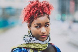 A young person with bright red curly hair is gazing toward the camera with a neutral expression. They are wearing large geometric earrings and a multicolored scarf, blending into the urban workforce. The background is blurred, suggesting an outdoor city setting.