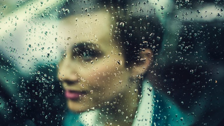 A woman with short hair, wearing a suit, is seen behind a rain-covered window. The blurred glass distorts her features, but her face is visible with raindrops in sharp focus. The background of greens and blues suggests an outdoor scene, blending the workforce imagery into a poignant moment.