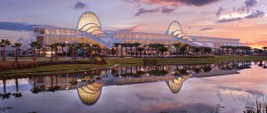 A modern building with arched glass rooftops is illuminated at sunset, reflected in a calm lake in the foreground, with palm trees and a colorful sky in the background.