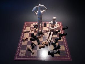 Two wooden artist mannequins on a chessboard set the scene. One stands with hands on its head, appearing frustrated, while the other sits, holding a chess piece. Scattered white and black pieces surround them in various positions on the board.