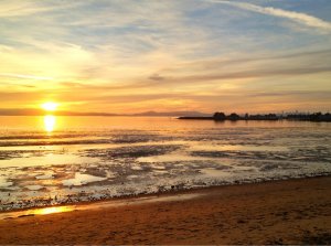 A serene beach scene during sunset. The sky is painted with warm shades of orange and yellow, reflecting on the calm water. The horizon features distant mountains and a mixture of clouds. The beach is peaceful, with shallow waves gently touching the shore.