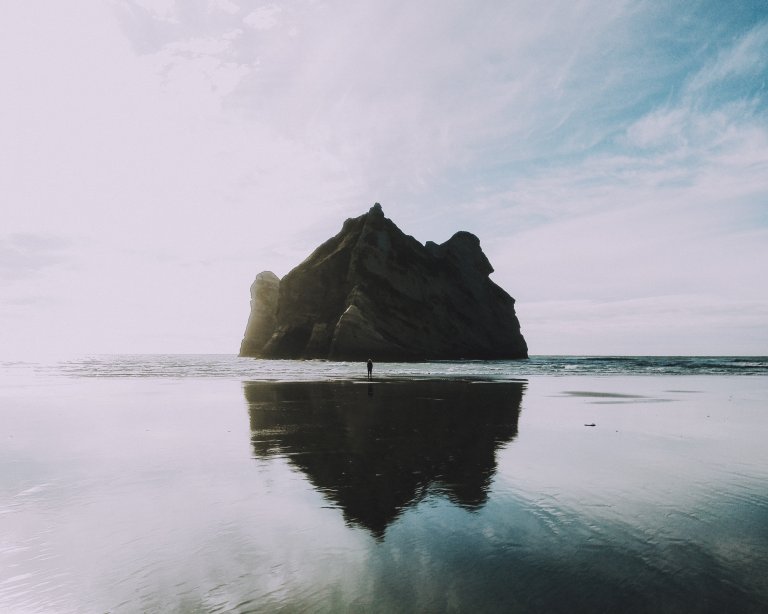 A large rock formation stands in the shallow waters of a beach under a partly cloudy sky. Its silhouette is mirrored on the wet sand below. A solitary figure is seen near the base of the rock, contemplating their decisions, adding a sense of scale to the vast landscape.