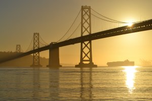 A bridge spans across the water during sunset, with the sun partially hidden behind the bridge and casting a golden reflection on the water. In the background, a container ship is seen moving towards the horizon. The scene is enveloped in a warm, golden haze.
