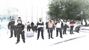 A group of construction workers stand and sit in front of an industrial building under construction. Clad in safety vests and hard hats, some are on their phones while others stand with crossed arms or engage in conversation. The scene captures the succession of daily routines, with trees visible in the background.