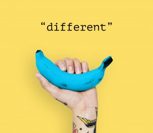 A hand with tattoos holds a blue banana against a yellow background. Above the banana, the word "different" is written in black text, symbolizing how successful people often embrace uniqueness and stand out.