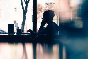 Silhouette of a person sitting in front of a laptop in a cafe, wearing a hoodie and beanie, talking on the phone. The background shows an outside view through large windows, with reflections and blurred effects creating a calm, focused atmosphere ideal for professional development.