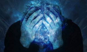 A person with disheveled hair holds their head in both hands, appearing distressed or overwhelmed. The position of their hands emphasizes their anguish. The image is overlaid with a blue, icy texture, adding a surreal and intense atmosphere. The background is dark and indistinct.