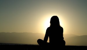 Silhouette of a person sitting cross-legged on the ground, facing a stunning sunrise or sunset with mountains and a calm body of water in the background. The sun's glow creates a serene and contemplative atmosphere, inviting you to lose yourself in the moment.