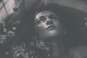 Black and white photograph of a person, perhaps yourself, partially submerged in water with face upward and closed eyes, creating a serene and introspective mood. Surrounded by floating flowers and petals, their face reflects light, highlighting a contemplative expression.