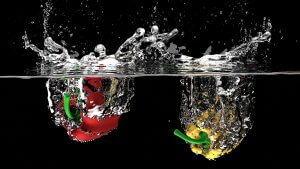 A red bell pepper and a yellow bell pepper partially submerged in water against a black background create splashes and bubbles as they hit the surface, their stems visible. The water's clarity contrasts with the vibrant colors of the peppers, echoing the vitality central to worksite wellness programs.