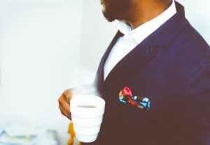 A man in a navy blue suit with a colorful pocket square holds a white cup of coffee. The focus is on the upper part of his body, projecting a sense of safe elegance as he looks away from the camera. The background is subtly blurred.
