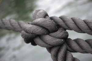 Close-up of a thick black rope tied in a knot, its weathered texture in sharp focus. The knot is tightly wound and prominently positioned at the center of the image, contrasted against a blurred, water-like background.