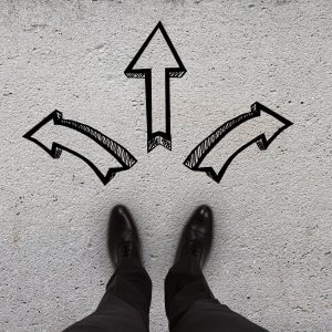A person wearing black dress shoes stands on a concrete surface, facing three painted arrows. One arrow points left, one points right, and the largest arrow points straight ahead, academically indicating different possible directions to take.