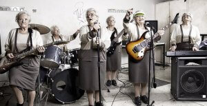 Six elderly women defy ageism in a band, each playing different instruments. One sings into a microphone, one stands at a drum set, one plays bass guitar, one raises a hand with an electric guitar, one is at a keyboard, and one stands proudly holding an electric guitar.