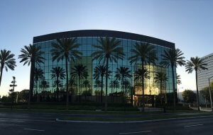 A modern glass office building with palm trees lining the front is reflected in the windows. The sky is clear, and the sun is either rising or setting, creating a warm, golden glow on the building and the trees. The scene is urban with minimal traffic.
