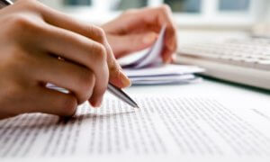 A close-up of a person's hand holding a pen, meticulously writing on what appears to be a cover letter. The other hand grasps several papers. In the background, a white keyboard and a blurred window hint at a busy workspace environment.