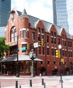 A historic red brick building with arched windows and a turret stands at a corner of a downtown intersection. Modern skyscrapers surround it in the background. A vintage clock and street signs are in front, marking Main Street and 4th Street.