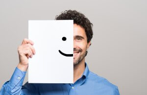 A person with curly hair and a blue shirt, likely one of the employees, holds a piece of paper in front of their face. The paper has half of a simple smiley face drawn on it, humorously aligning with the person's real smile to create an illusion of a complete face. The background is plain gray.