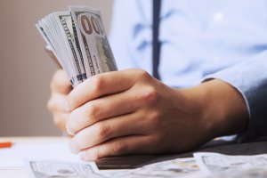 A person wearing a light blue shirt and dark tie is holding a stack of U.S. $100 bills in their hands, suggesting they are financially fit. The table in front of them has more scattered bills. The background is out of focus.