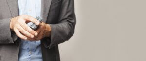 A close-up of a person in a gray blazer and light blue shirt holding a stack of cash, with only the hands and part of the torso visible. The background is plain and light-colored, projecting an image of being financially fit.