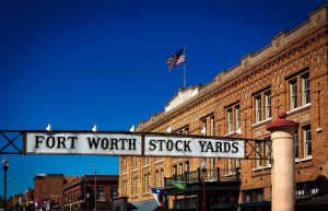 A sign reading "Fort Worth Stock Yards" hangs over a street lined with historic brick buildings. The American flag waves atop one building against a clear blue sky.