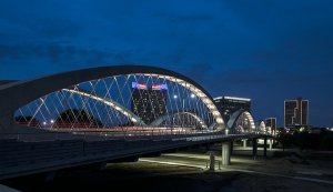 A modern, arched bridge illuminated with lights spans across a waterway at night. The bridge features a sleek design with crisscrossing cables. In the background, several high-rise buildings lit up with lights can be seen against a dark blue sky.
