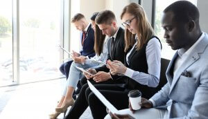 A group of five people, dressed in business attire, sit in a row on chairs, each focused on their respective documents and digital devices. Despite the diverse ages represented, there's no hint of ageism. The background showcases large windows with an expansive view outside. One individual holds a cup of coffee.