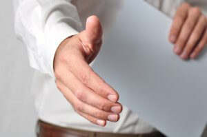 A close-up of a person extending their right hand as if offering a handshake, symbolizing unity and combating issues like ageism. The individual is wearing a white long-sleeve shirt, and the background remains unobtrusive to keep the focus on this welcoming gesture.
