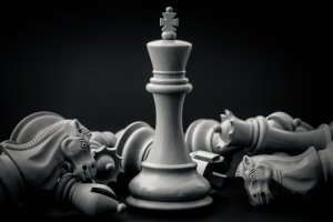A central wooden chess king stands upright, embodying leadership, surrounded by toppled knights, rooks, and pawns against a dark background. The black and white image emphasizes the texture and craftsmanship of the chess pieces.