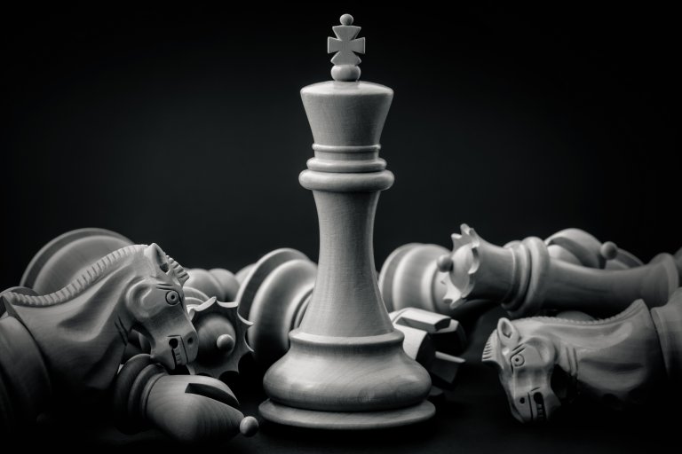 A central wooden chess king stands upright, embodying leadership, surrounded by toppled knights, rooks, and pawns against a dark background. The black and white image emphasizes the texture and craftsmanship of the chess pieces.