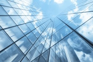 A view looking up at the reflective glass facade of a skyscraper, capturing the blue sky and clouds mirrored on its surface. The architecture creates a symmetrical and abstract pattern, emphasizing the building’s height and modern design, symbolizing leadership in urban development.
