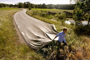 A person of strong character appears to be pulling back the pavement of a winding road like a sheet, revealing the ground beneath. The scene is set in a rural area with lush greenery, a tranquil pond, and trees in the background, under a partly cloudy sky.