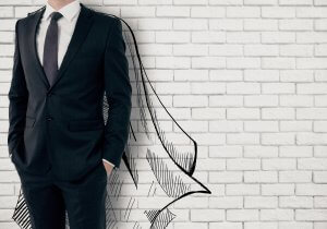 A man in a suit stands against a white brick wall with hands in his pockets. A superimposed black and white drawing of a superhero cape is outlined on the wall, giving the illusion that he is wearing it, embodying leadership.