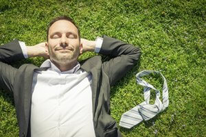 A man in a suit and white shirt lies on green grass with his eyes closed and hands behind his head, looking relaxed. A striped tie is lying beside him. The scene suggests he is taking a break from the work day or enjoying a moment of peace outdoors.